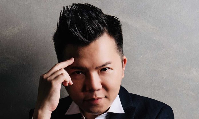 Closeup photo of Dato' Anthony Cheng in a collared shirt in a 'thinking' pose with a finger pointed to his head, against a gray gradient background