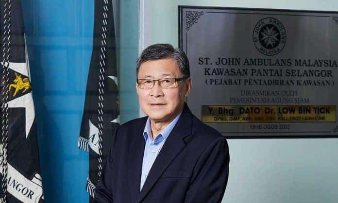 Medium closeup photo of Dato' Yeo Kim Thong in a light blue dress shirt and black suit at St John Ambulance Malaysia Kawasan Pantai Selangor Area HQ wtih an award (of Dato' Dr. Low Bin Tick) and flags in the background