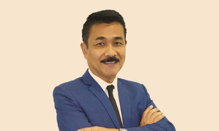 Bust portrait photo of Datuk Zakaria in a bllue suit and tie with his arms folded against a tan background