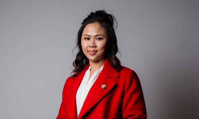 Medium closeup photo of Dr Quirix Tan in a white top and red suit with a brooch, against a gray background