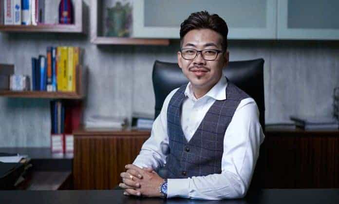 Medium closeup pohoto of Edison Ser in a white dress shirt and gray vest sitting at his office desk with bookshelves and cabinets in the background