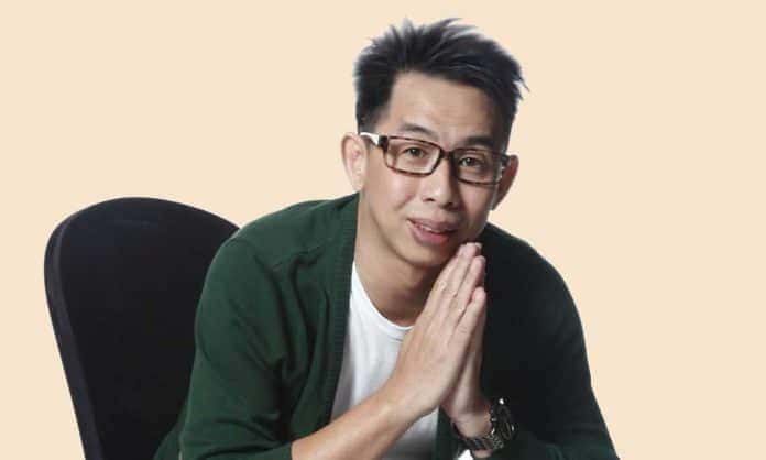 Photo of Ernest Kok in a white shirt and dark green jacket sitting in chair leaning forward with palms together with tan background