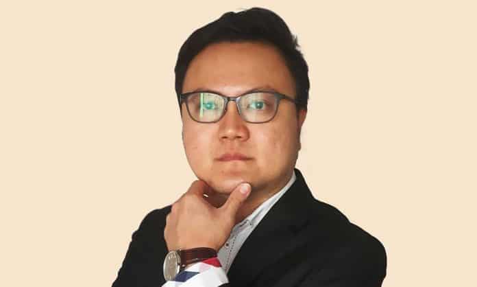 Bust portrait photo of Ian Lau in a black suit, with a watch and cloth wrapped around his left hand held up to rub his chin, posed against a tan backgrounf
