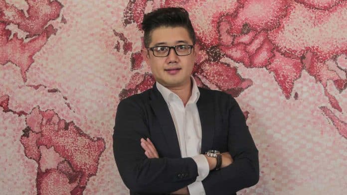 Half-body photo of Jackal Chua in a white dress shirt and black suit with pink wallpaper depicting the world map in the background