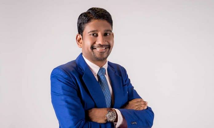 Medium closeup photo of K. Anandraj in a blue suit and tie with his arms folded against an off-white background