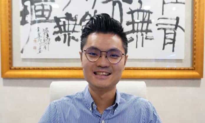 Bust portrait photo of Ken Teo in a striped blue shirt sitting in a white chair with framed Chinese calligraphy in the background