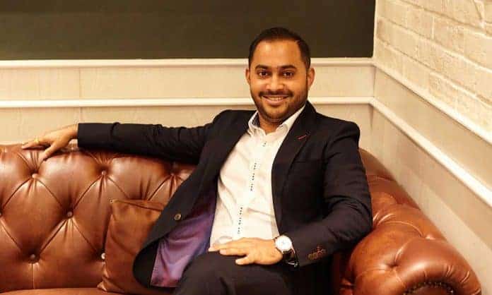 Sri Vadivale in a white dress shirt and black suit reclining on a brown couch with his arm rested on top