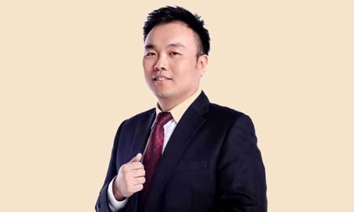 Medium closeup photo of Tee Kiat Siong in black suit and red tie with tan background