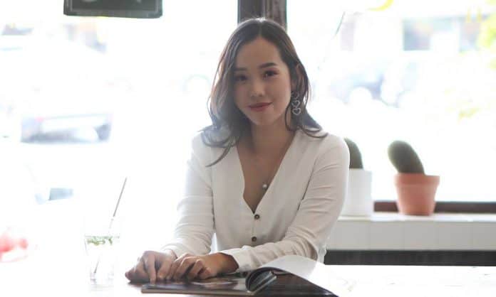 Medium closeup photo of Wendy Tan in a long-sleeve white top at a white table with an open book and beverage against a bright blurred background