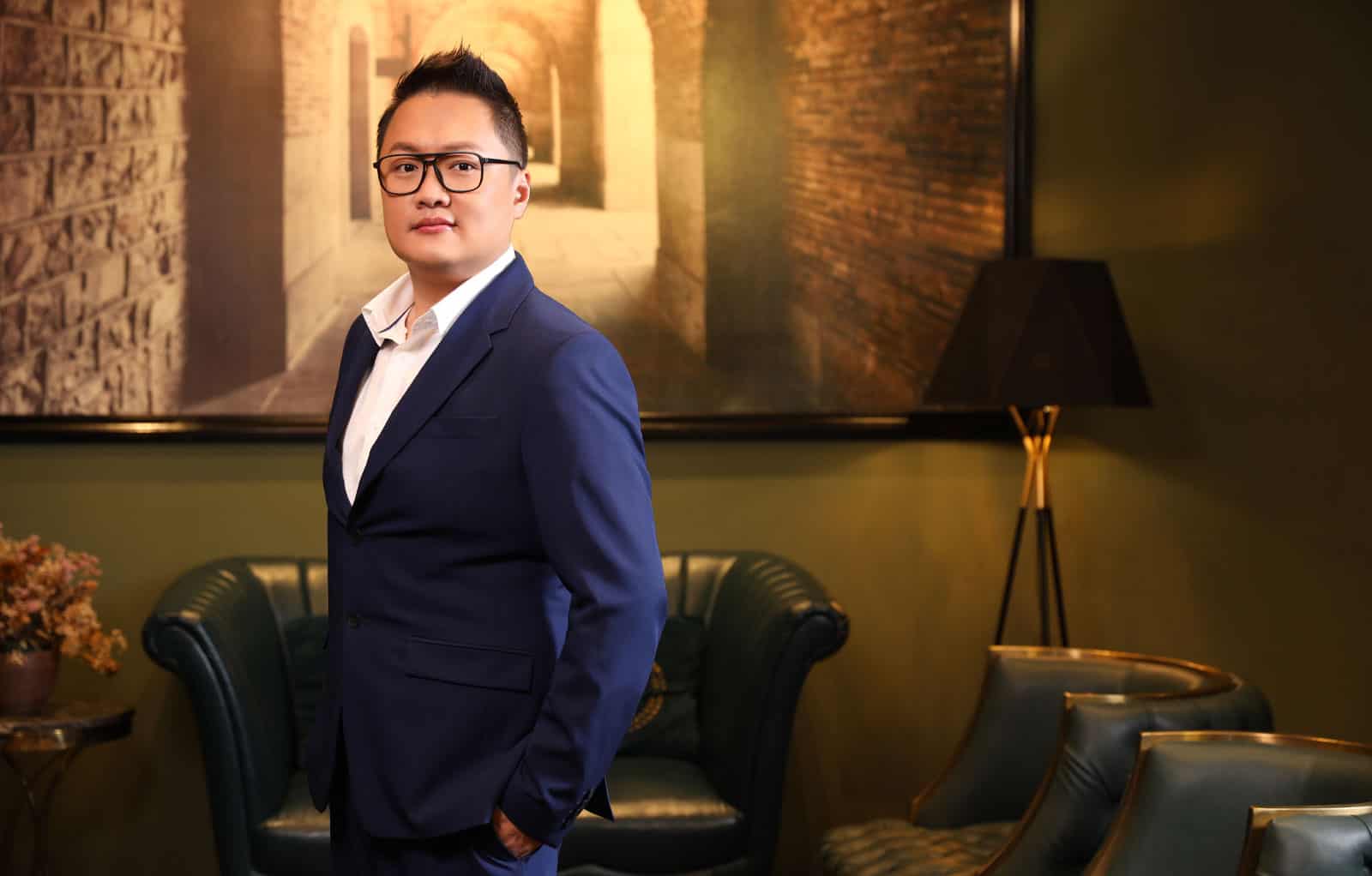 Half-body portait photo of Adrian Yee in a dark blue suit within a warmly lit room with furniture and a large framed image