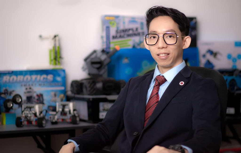 Photo of Aljay Ng in a black suit and red tie, seated,  with a collection of robots in the background