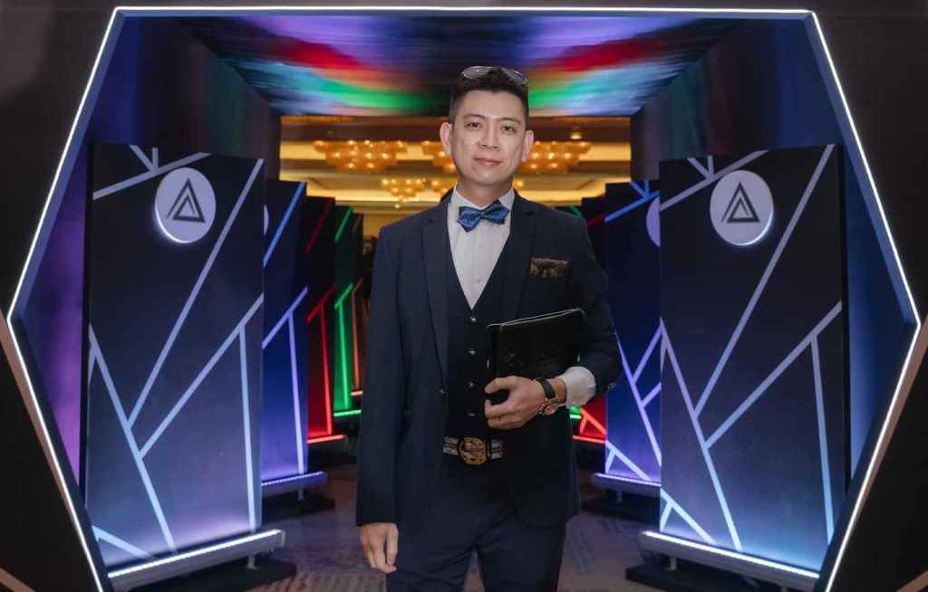 Photo of Ayden Mok Chee Chuan in a blue suit and bowtie, standing in front of displays of award categories in Leadership Excellence Awards