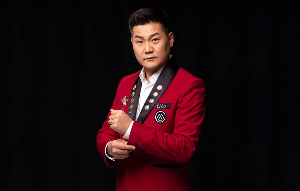 Half-body portrait photo of Bayarbold Enkhkhishig in a white shirt and red suit with black lapels adorned with gold and silver pins; standing in front of a black background, adjusting his cuffs
