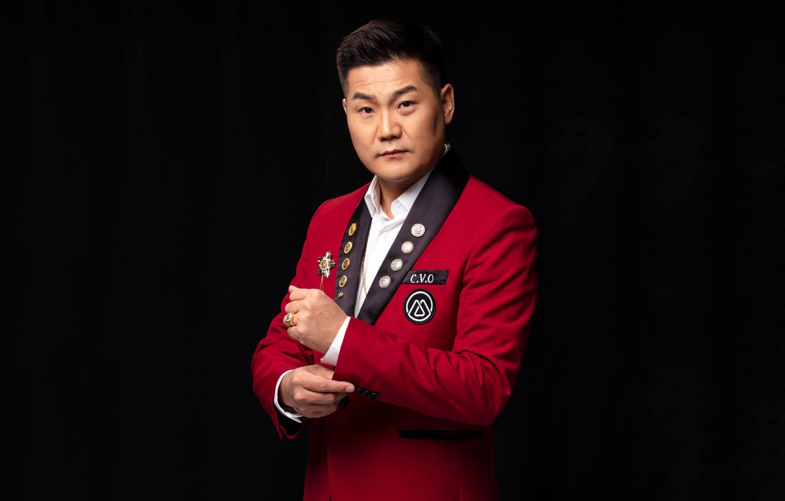 Half-body portrait photo of Bayarbold Enkhkhishig in a white shirt and red suit with black lapels adorned with gold and silver pins; standing in front of a black background, adjusting his cuffs