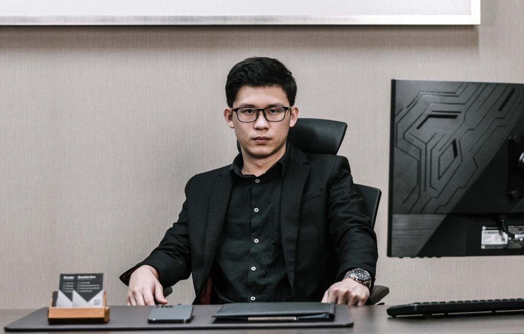 Photo of Benedick Chen Hau Cher in black shirt and suit at his office desk with a light gray background