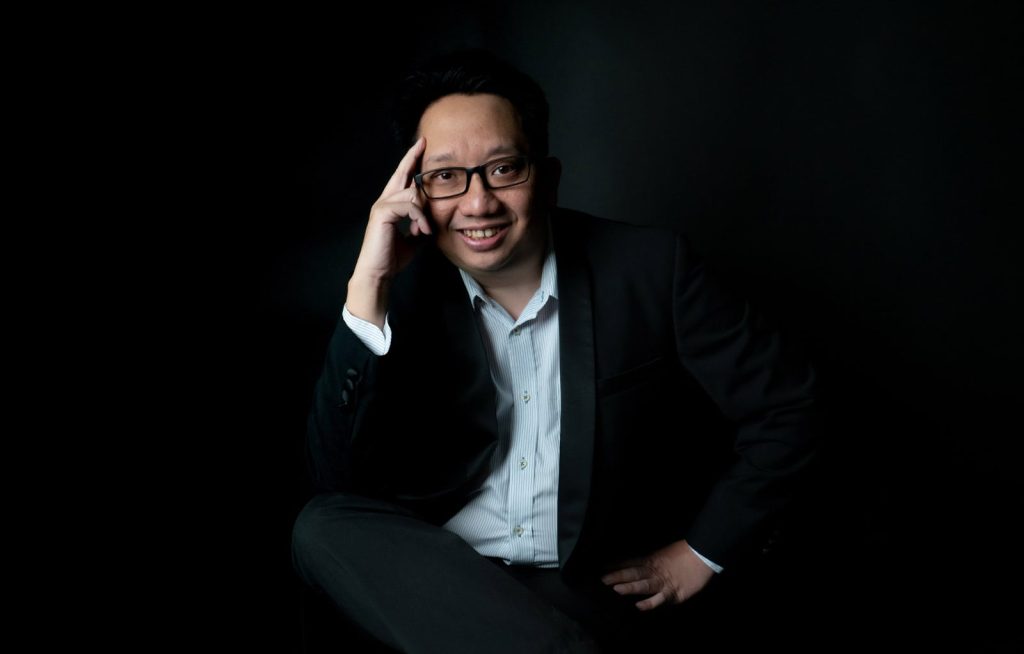 Portrait photo of Charles Gregory in a white shirt and black suit, one hand held up to his temple, and the other on his waist, seated with legs crossed, against a black background
