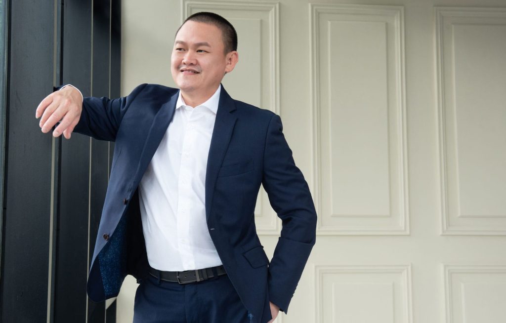Half-body portrait photo of Chen Ling Choy in white dress shirt and black suit leaning against wall