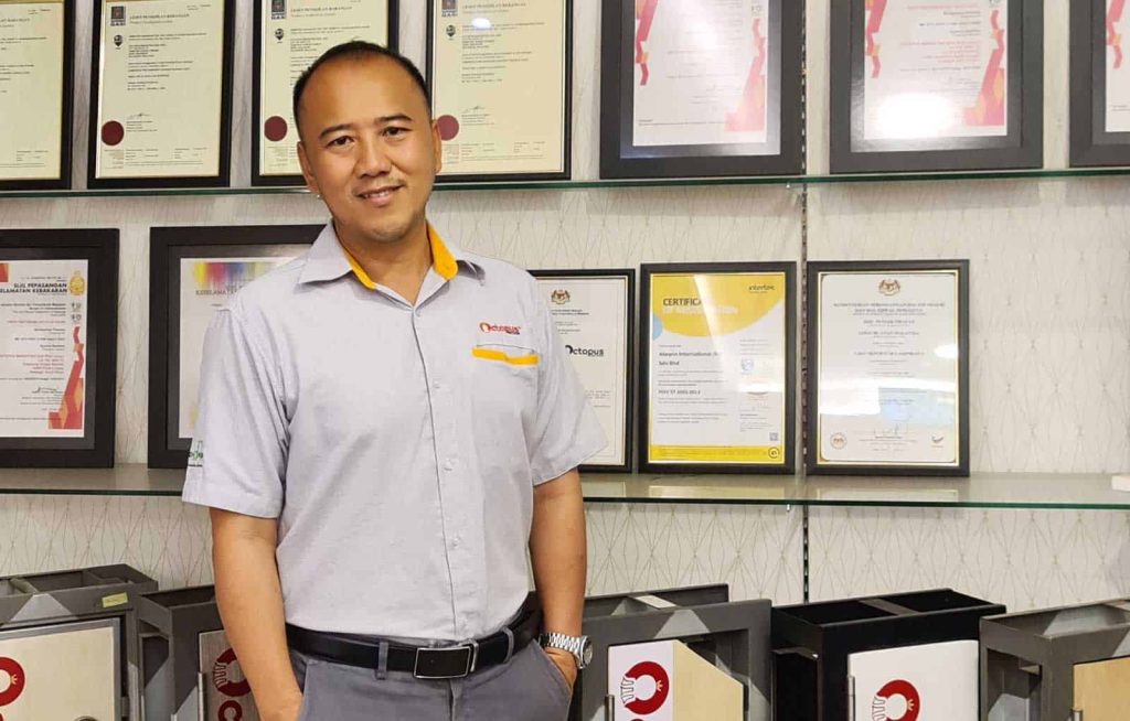 Half-body portrait photo of Daniel Loke in the company uniform, with hands in pockets, standing in front of a wall of certificates