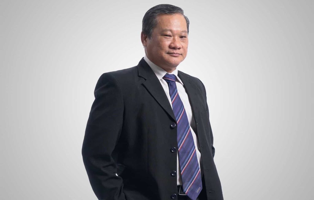 Half-body portrait photo of Dato’ Ooi Win Juat in a white shirt, black shirt and striped purple tie; standing with hands in pockets in front of a white background