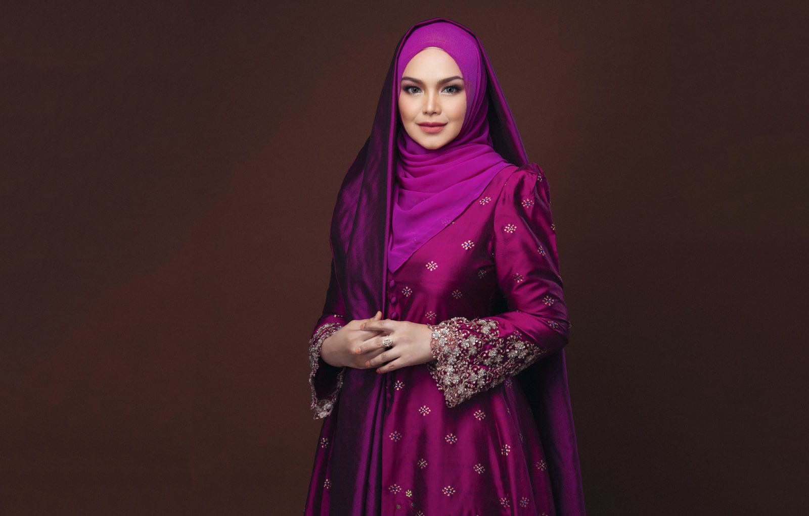 Half-body portrait of Dato' Sri Siti Nurhaliza Binti Tarudin in magenta dress and headscarf, hands held together with a dark brown background
