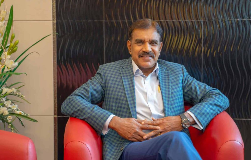 Seated portrait of Datuk Dr Selvam Rengasamy in a checkered blue-green suit, seated in a red chair with wavy back pattern on the wall and a plant in the background