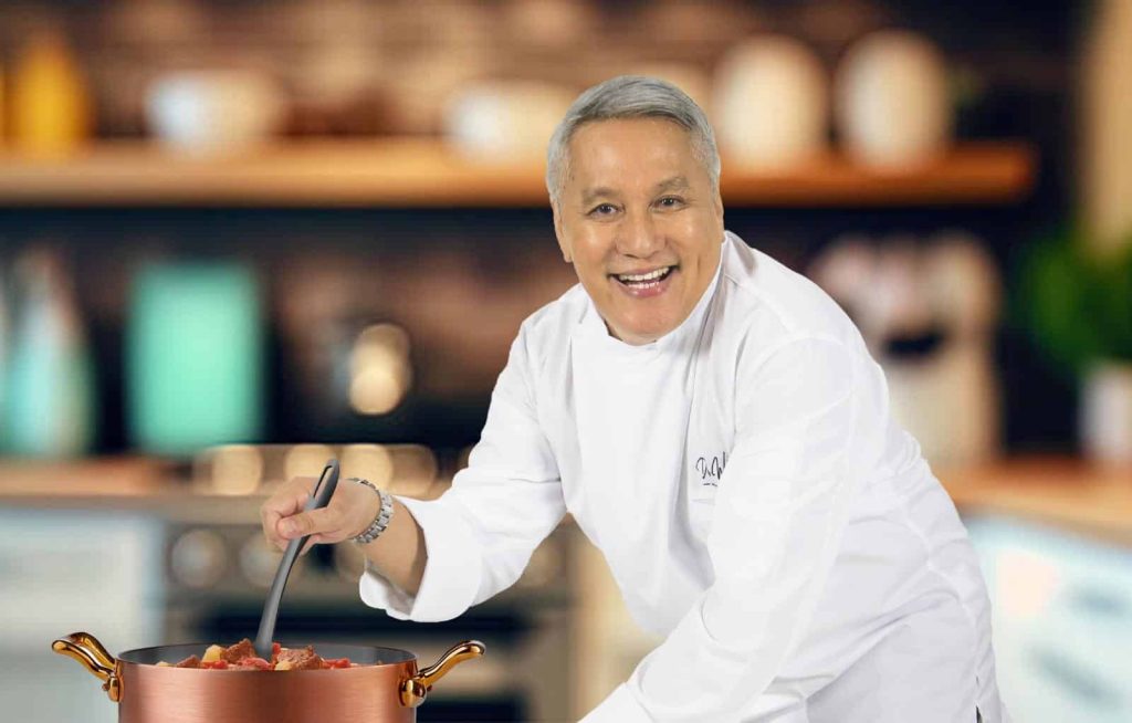 Portrait of Datuk Redzuawan bin Ismail in a chef uniform smimling and cooking - bending towards a cooking pot with a ladle in one hand