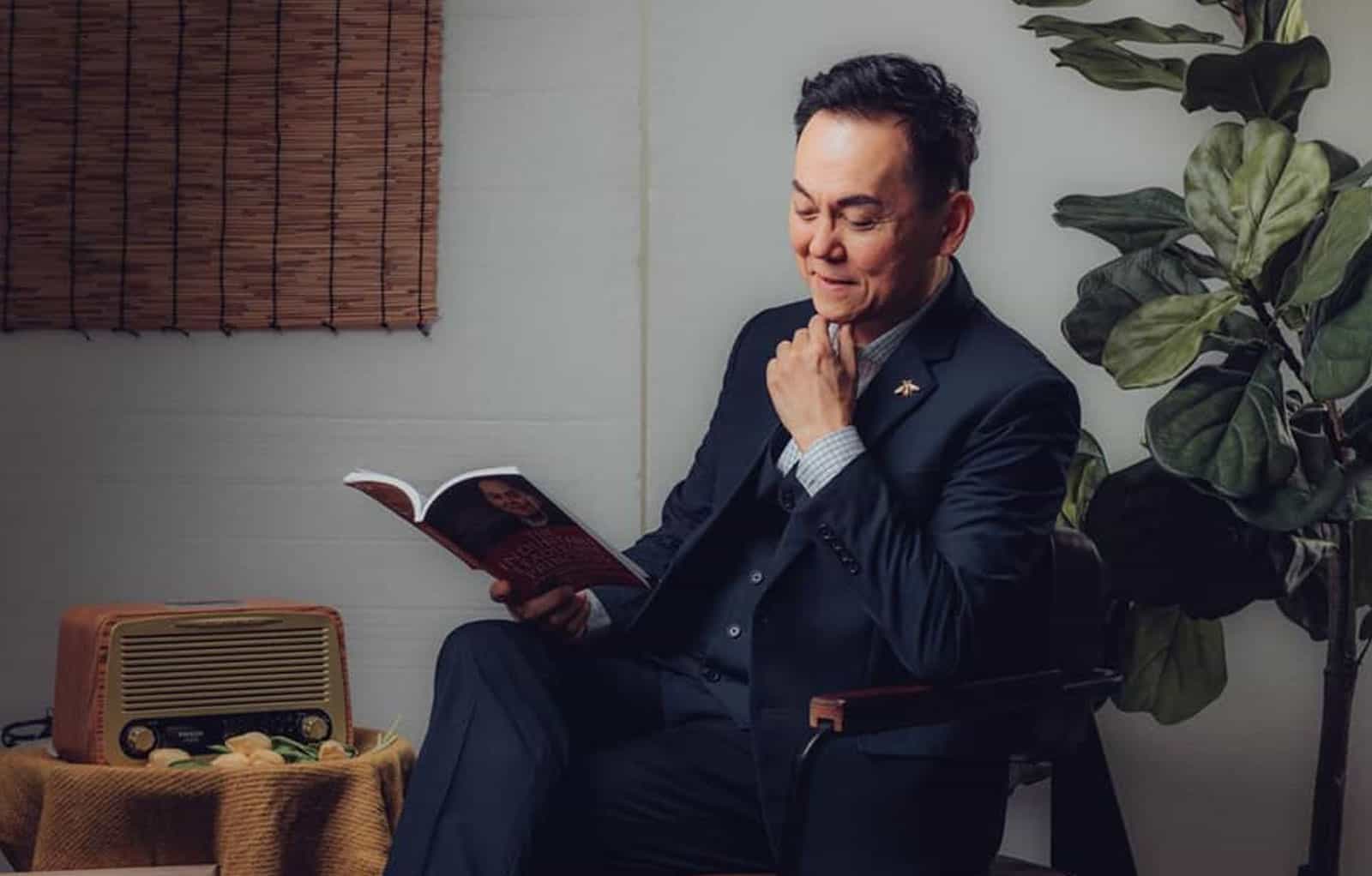 Portrait photo of Dr. Gilbert Ng Chong Kwee in a black vest and suit, sitting in a chair while reading and rubbning his chin, with a plant, drapery and an old school radio in the ground =