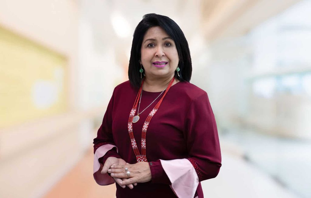 Half-body portrait photo of Dr Kuldip Kaur Prem Singh in a magenta dress with big light pink cuffs; accessorized with a long red traditional thread necklace, a silver pendant, a ring and green earings; holding her palms together; while standing in front of a blurred background of a room