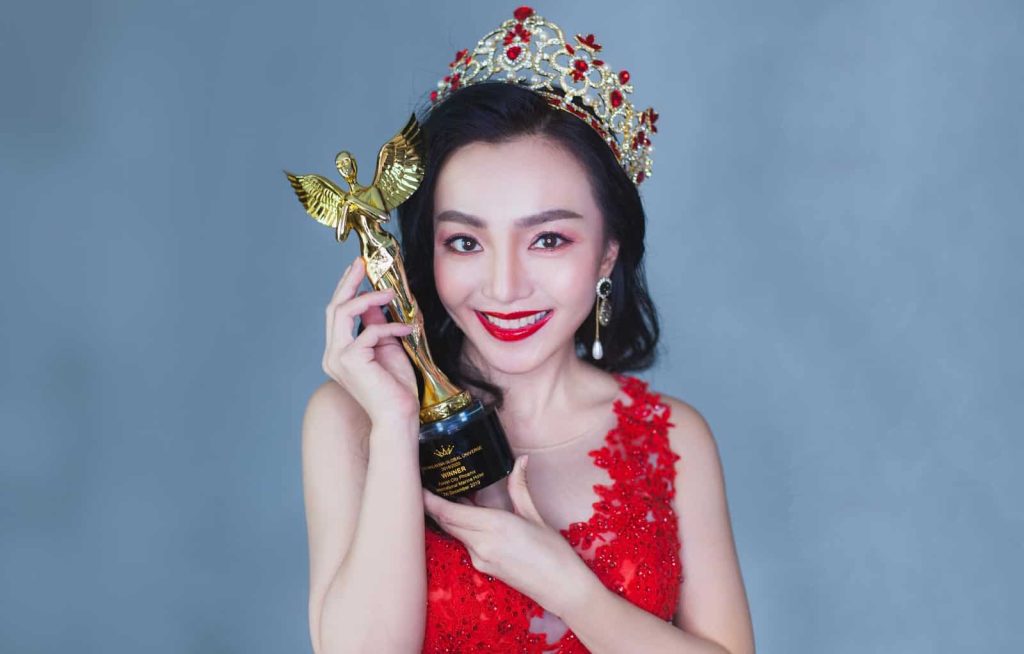Bust portrait of Dr Shana Yong in a crown and red dress holding up a golden trophy depicting a humanoid with wings