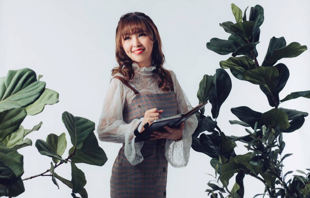 Portrait photo of Elaine Gan Lee Chian in dress on top of a sheer fabric top holding up and writing on a book surrounded by plants with a white background