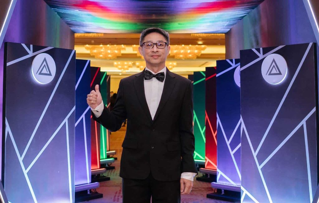 Photo of Eric Lim in a black suit and bowtie standing in front of award category displays at Leadership Excellence Awards