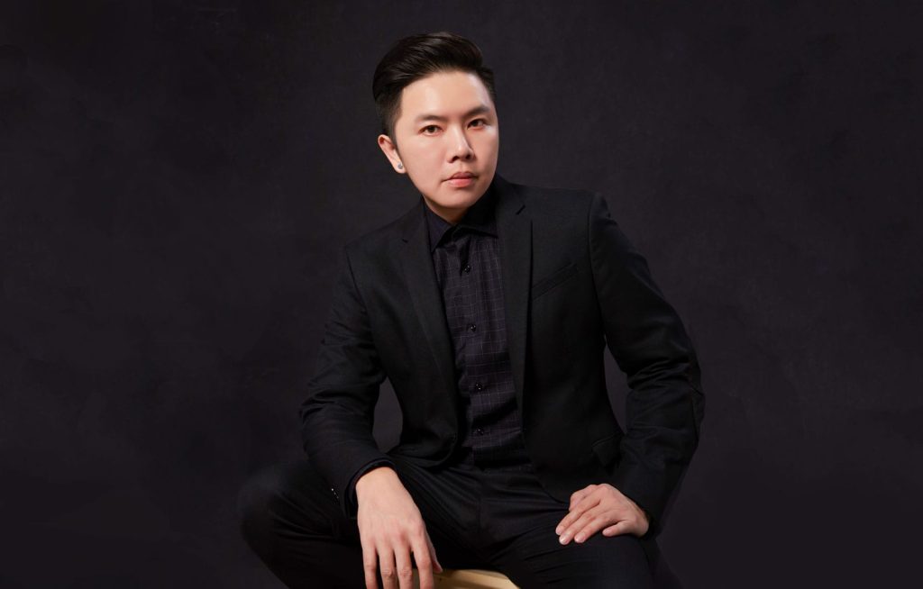 Seated portrait of Fui Chong in a black suit sitting on a stool in front of a black bacgrounf