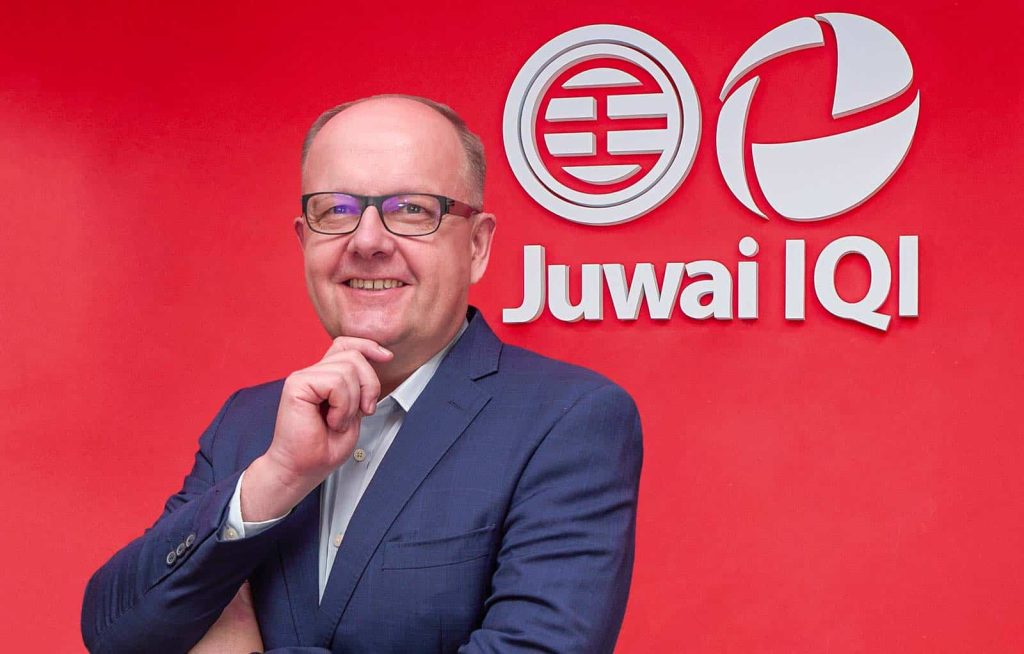 Medium closeup portrait photo of George Chmiel in a white shirt and gray suit, posed with a hand held up to his chin; standing in front of a red wall with the Juwai IQI logo printed