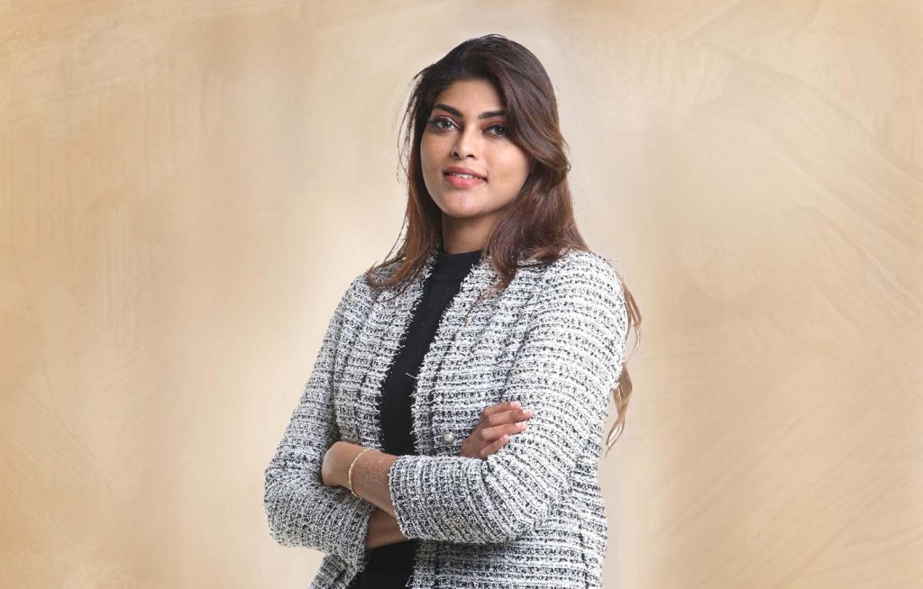 Portrait photo of Hemala AP Sivam in a black top and light gray thread jacket, posing with arms folded in front of a textured cream background
