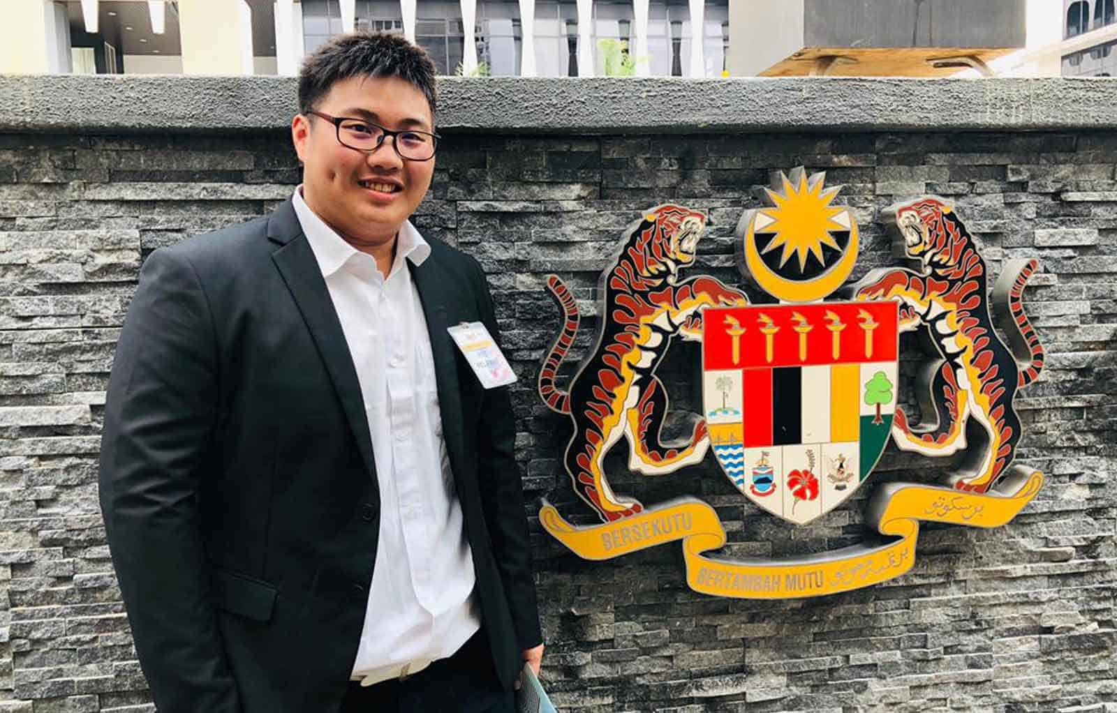 Half-body photo of Jayson Ho Jie An in a white shirt and black suit with the Malaysian a display of the Malaysian coat o0f arms in the background