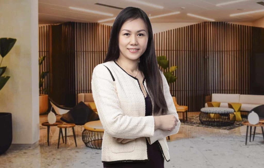 Half-body portrait photo of Jeanie Ho in a black dress and white top in a warmly lit room furnished with chairs, tables, and plants