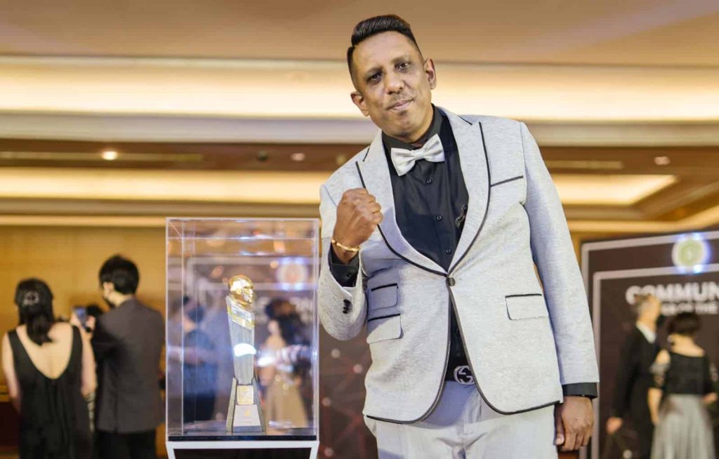 Photo of Jeevanand Muniandy in a light gray suit and bowtie, standing next to a LEA trophy with other attendees of the event milling about in the background