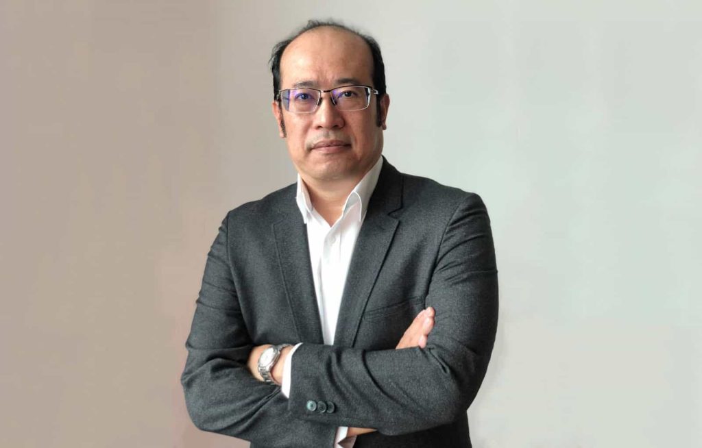 Half-body portrait photo of Kenneth Kee in a black suit, with arms folded, against an off-white gradient in the background