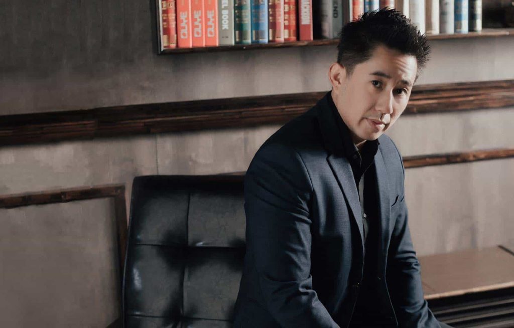 Portrait photo of Lathen Loh in a black suit seated on a couch surrounded by dark brown walls a bookshelf in the background