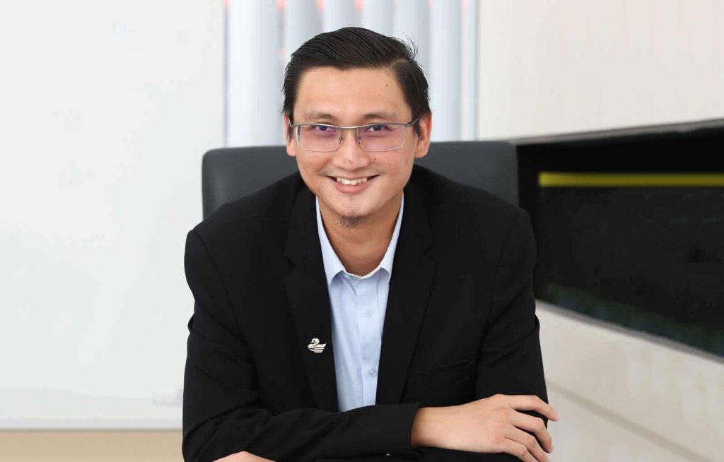Closeup portrait of Mohd Hafizuddin Hiew bin Abdullah in a black suit, leaning forward with arms folded, seated at an office desk