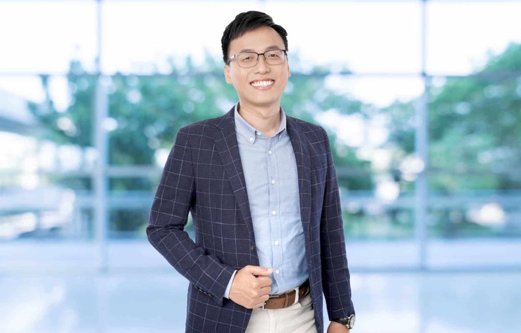 Half-body portrait photo of Ong Hang Ping in dress shirt and suit against a blurred background with trees behind windows