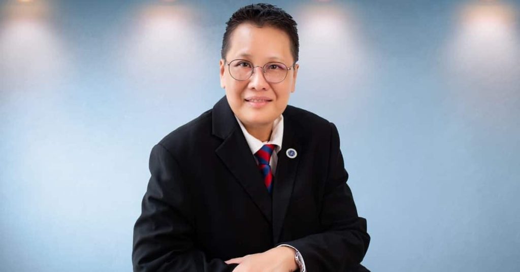 Bust portrait photo of Ruth Chai in a blue shirt and black suit with hands clasped, and a gray background with warm lights