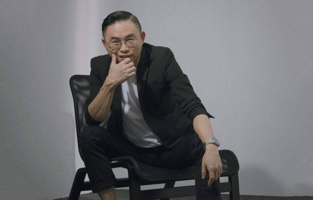 Seated portrait photo of Ryon Leee in a white shirt and black suit seated on a chair, with his hand rested near face, against a white wall