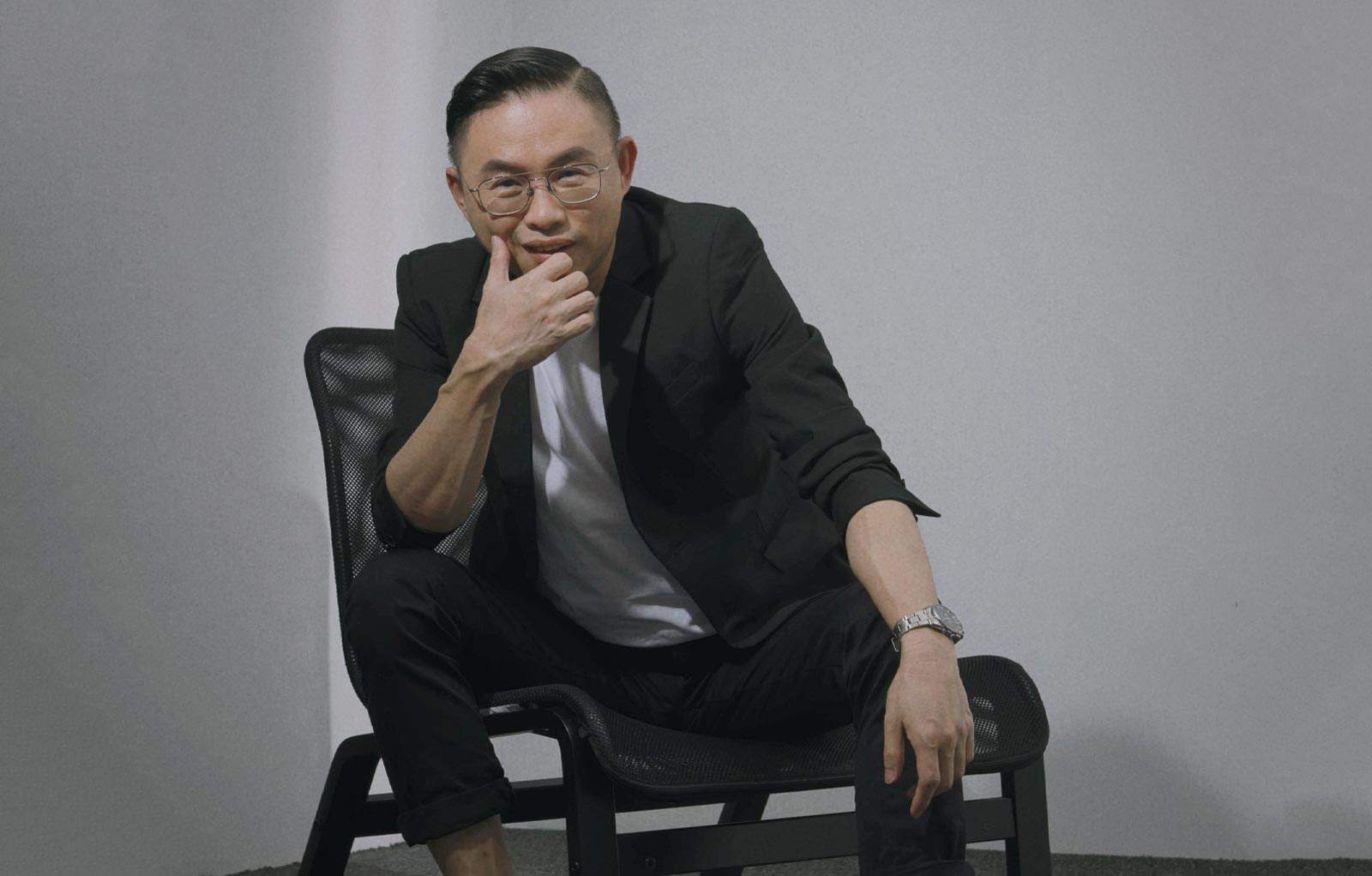 Seated portrait photo of Ryon Leee in a white shirt and black suit seated on a chair, with his hand rested near face, against a white wall