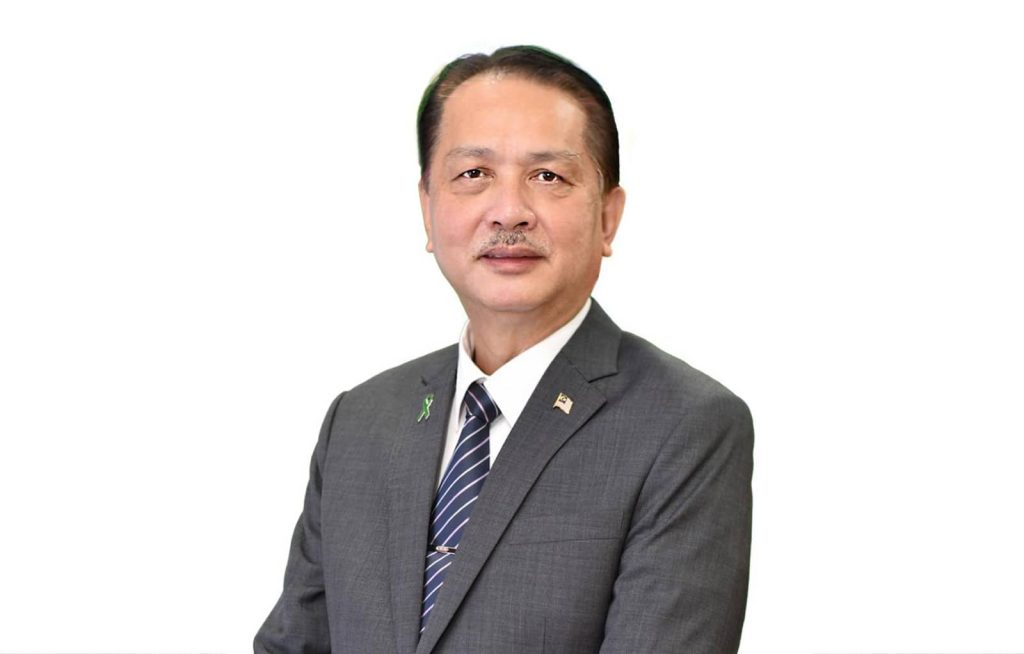 Bust portrait photo of Tan Sri Datuk Seri Dr Noor Hisham Bin Abdulla in a white shirt, gray suit and silver-srtiped blue tie, with pins on lapels; posed in front of a white background