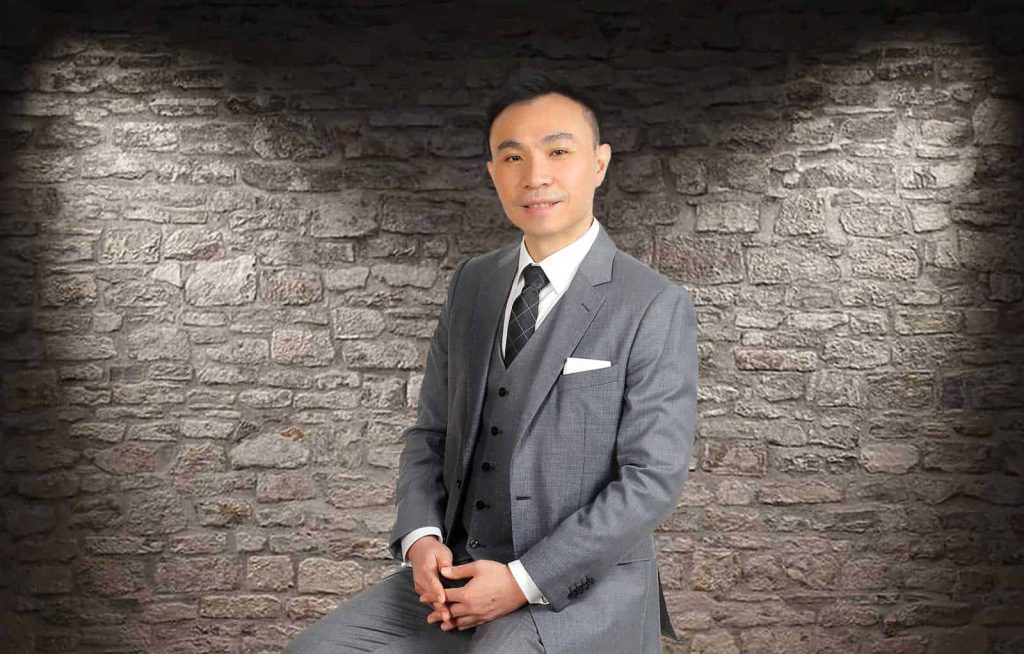 Half-body portrait of Terence Teoh Wee Khang in a gray suit and black tie, sitting with hands sclasped together, in front of a gray stone wall
