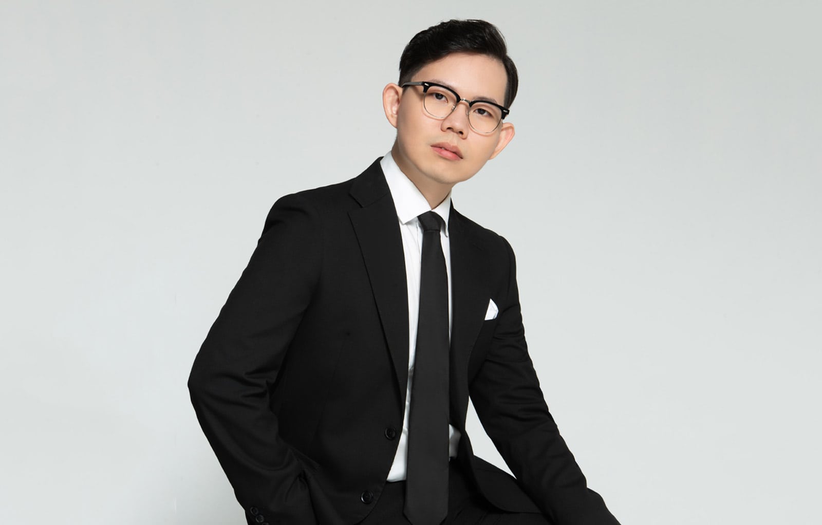 Portrait photo of Wilson Tay Kah Hoe in a black suit and tie, sitting cross-legged, with a white background