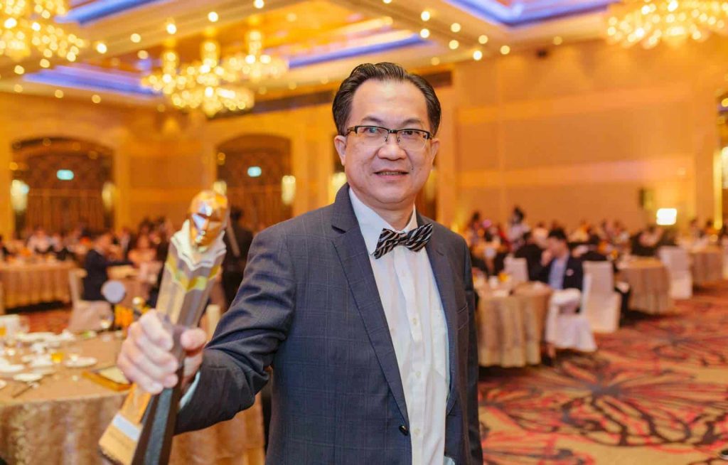 Photo of Yeow Liang Ming in a gray suit and striped bowtie at Leadership Excellence Awards, in a warmly lit ballroom, holding up a trophy